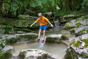 Young boy in orange shirt jumping over clear stream between mossy rocks in lush green forest with small waterfall, capturing dynamic moment of outdoor adventure.