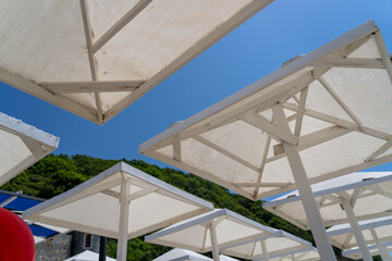 Upward view of numerous white beach umbrellas and their wooden frames against clear blue sky, overlooking verdant hillside on bright sunny day at resort