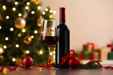 Fototapeta premium Glass of red wine, bottle and Christmas decor on wooden table against background with blurred lights