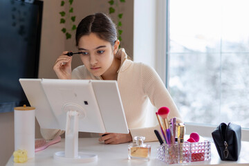 Teen girl applying mascara at makeup table. A focused young girl applies mascara in front of a lit vanity mirror, surrounded by makeup tools and natural daylight from a window.