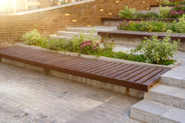 Sunny urban terraced garden with modern wooden slat benches, lush green and pink flowering plants in concrete beds, and brick wall architecture.