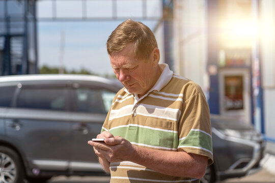Focused senior man in striped polo shirt checking his smartphone outdoors on bright day, with blurred cars and building structures in background.
