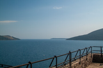 Expansive coastal panorama featuring tranquil blue sea stretching towards distant mountains under clear sky, viewed from stone railing overlook on bright day.