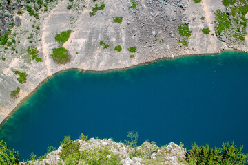 Overhead shot of pristine deep blue lake with clear turquoise edges, surrounded by rugged, arid landscape featuring rocky slopes and pockets of green vegetation