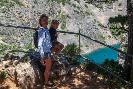 Woman and young girl pause on cliffside path, overlooking vibrant turquoise lake nestled among rugged mountains and steep rocky slopes. - Powered by Adobe