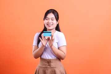 Naklejka premium A smiling young woman holds a credit card with her two hands, happy for the new financial possibilities, isolated on orange background.