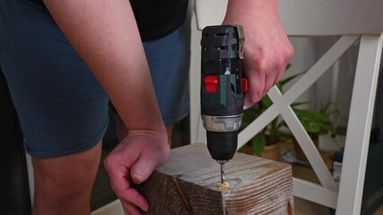 Man drilling hole into thick wooden beam block holding cordless drill driver with both hands at home interior. Concept of DIY woodworking project, home carpentry, power tool usage and handyman work.