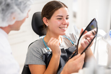 In dentists office, patient sits in dental chair with mirror in hands, see reflection, enjoys result of dental treatment. Male doctor dentist satisfied with result of prosthetics of girl patients