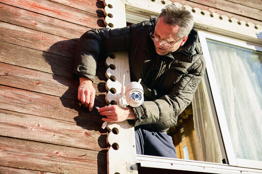 Linking technician connects cable to surveillance camera fixed on exterior wall of countryside home, directing wire to port during setup.