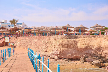 Beachfront scene featuring wooden pier leading to sandy shore, surrounded by straw umbrellas and rocky coastline, creating a vibrant summer vacation atmosphere
