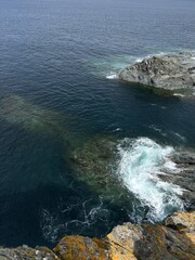 Top view of clear turquoise ocean water with visible underwater rocks and a rugged coastal cliff....