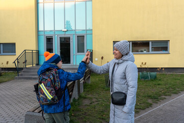 Smiling mother and son giving high five near school building entrance on cold autumn day in winter jackets. Concept of school achievement celebration, parent support, family bonding and motivation. 