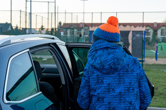 Boy in blue winter jacket and knitted hat with pompom standing near open car door looking at playground in suburban area. Concept of arrival for outdoor play, childhood recreation and family trip.