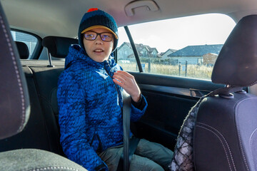 Young boy in blue winter jacket and knitted hat fastening seat belt in car back seat on sunny day. Concept of child safety, road trip preparation, responsible travel and family automobile journey.