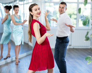 Obraz premium Enthusiastic young woman in vibrant red dress enjoying active dancing paired with male partner in formal wear in dance studio, practicing playful twist while female teacher observing from background