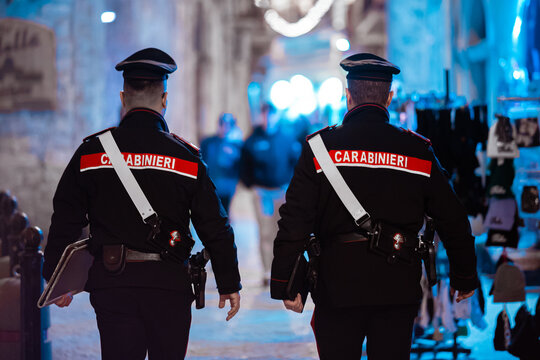 Rear view of two uniformed Italian Carabinieri police officers patrolling a dimly lit, atmospheric, historic street or alleyway in an Italian old town like Bari.