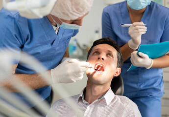 Elderly man dentist examining male patient, young female nurse taking notes in dental clinic