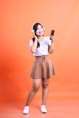 A joyful Asian girl is listening to music with headphones and a smartphone. Full shot. isolated on orange background.