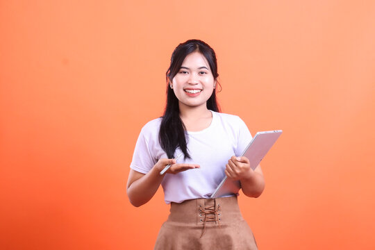 Smiling asian woman holding a tablet and pen with hand open in presenting gesture, ready to show some information, isolated on orange background
