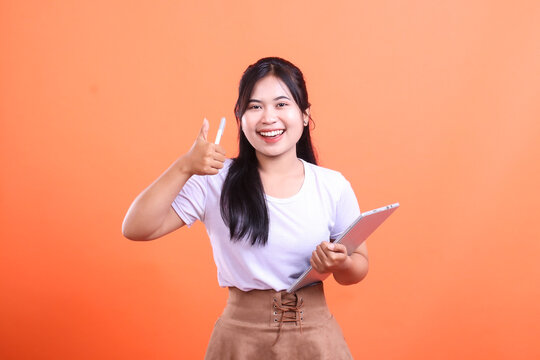Happy young woman giving a thumbs up while holding a digital tablet and smiling broadly to the camera isolated on orange background.