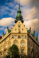 Obraz premium A grand historic building in Prague featuring a green copper spire, intricate facades, and colorful murals, capturing European architectural charm and cityscape ambiance.