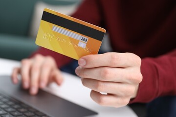 Young man making online payment with laptop and credit card at white table indoors, closeup