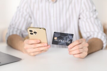 Young man making online payment with phone and credit card at white table indoors, closeup