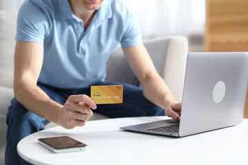 Young man making online payment with laptop and credit card at white table indoors, closeup