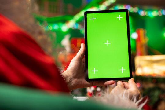 Man wearing Santa costume using mockup tablet in workshop to check messages from kids worldwide. Father Christmas enjoying winter holidays wishes sent by children via email using chroma key device