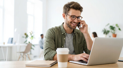 Smiling young businessman talking on the phone while working on his laptop in a bright, modern corporate office space.