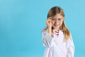 Smiling little girl in laboratory coat and glasses on light blue background, space for text. Child...