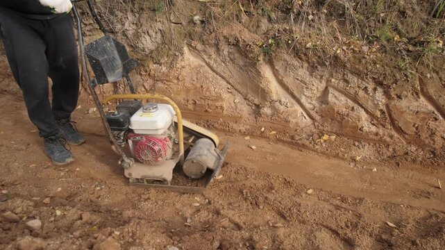 soil tamping with machine, operator uses compactor on trench soil, shallow trench compaction for utility pipes with equipment, closeup of compactor handling dry dust on grassy bank