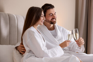 Happy couple in bathrobes with glasses of sparkling wine on bed indoors