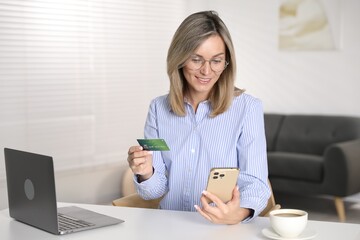 Woman making online payment with phone and credit card at white table indoors