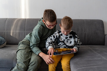 Two brothers sitting on grey sofa playing together on tablet with older boy helping younger sibling at home. Concept of siblings bonding, digital learning, screen time and childhood technology usage.