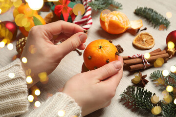 Woman decorating tangerine with cloves at light wooden table, closeup. Bokeh lights
