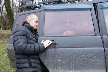 A middle-aged man with short hair is sitting in a car, focused on repairing a vehicle. There's a dog in the passenger seat.