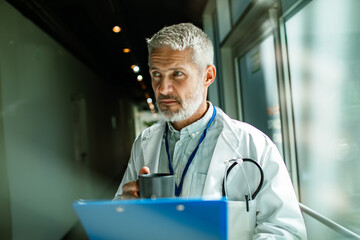 Mature male doctor with stethoscope drinking coffee in hospital corridor, thoughtful