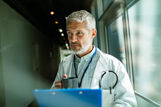 Mature male doctor with stethoscope drinking coffee in hospital corridor, thoughtful