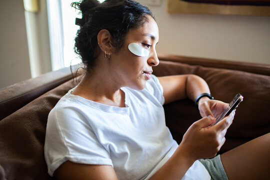 Young adult woman relaxed with eye patches using smartphone at home