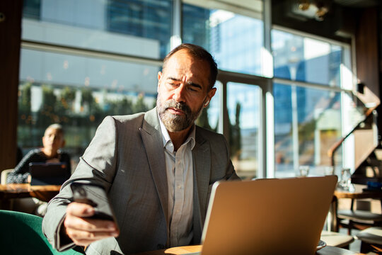 Mature man focused on phone and laptop in modern cafe