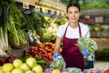Woman seller in a red apron holds a bag of edamame and puts it next to other vegetables and herbs. Supermarket employee is busy displaying products