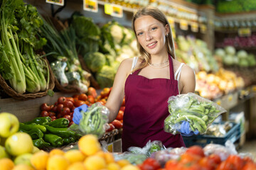 Girl puts ripe packaged green beans in pods in box on display case, arranges assortment in vegetable shop. Worker puts vegetables in pile pyramid. Employee reorganizes showcase.