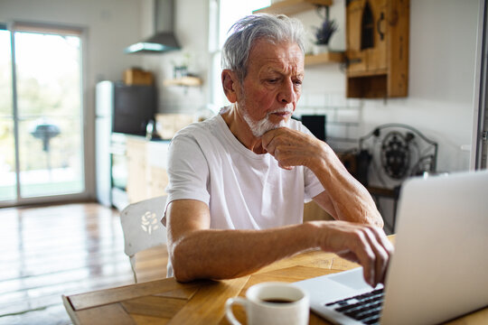 Senior man looking thoughtful using laptop at home kitchen
