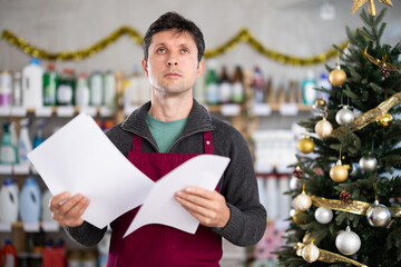 Anxious adult man seller with checklist in household chemicals store decorated for christmas