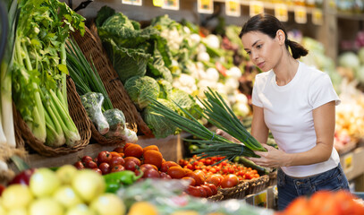 Girl client near vegetable stand in store choose bunch of green onion. Young woman shopping in vegetable store, customer takes out fruits from showcase, collects grocery basket..