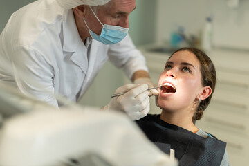 During reception, dental practice, mature male dentist doctor diagnoses teeth of patients girl, examines condition of oral cavity, gums, teeth. Doctor looking for bad tooth and source of pain
