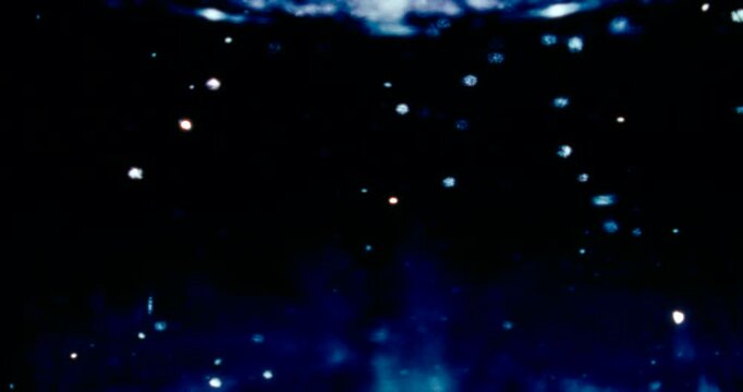 Particles float in water over a dark background, reflecting top light in green and blue hues, creating a glittering, halo-like, cosmic universe effect.