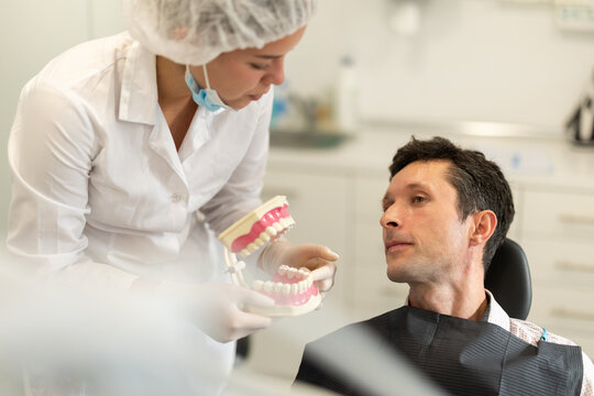 In dentists office, girl doctor conducts appointment and shows man patient plastic prop. Layout of jaws explains location of patients problem teeth using layout