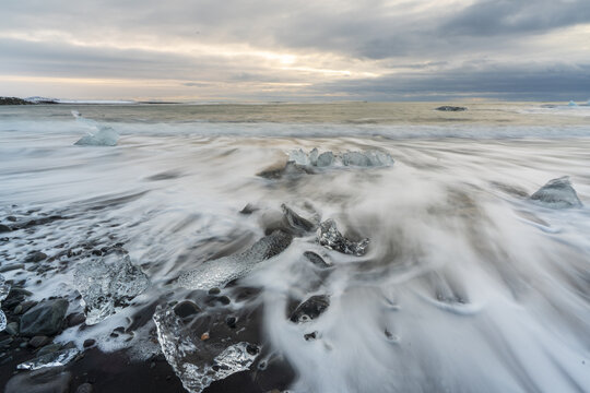 View of glacial ice fragments scattered across the black volcanic sand, as the tide ebbs and flows under a dramatic sky, Diamond Beach, Sveitarfelagid Hornafjordur, Iceland.
